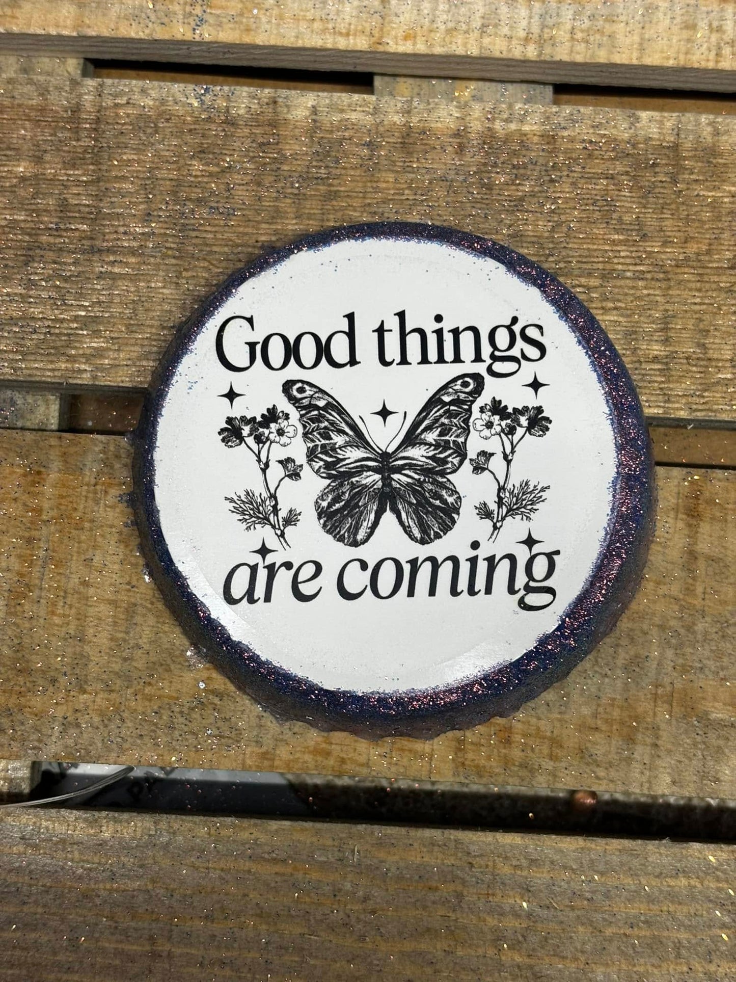 Round handmade freshie with black text that reads “Good things are coming,” centered around a butterfly and floral design on a wood background.