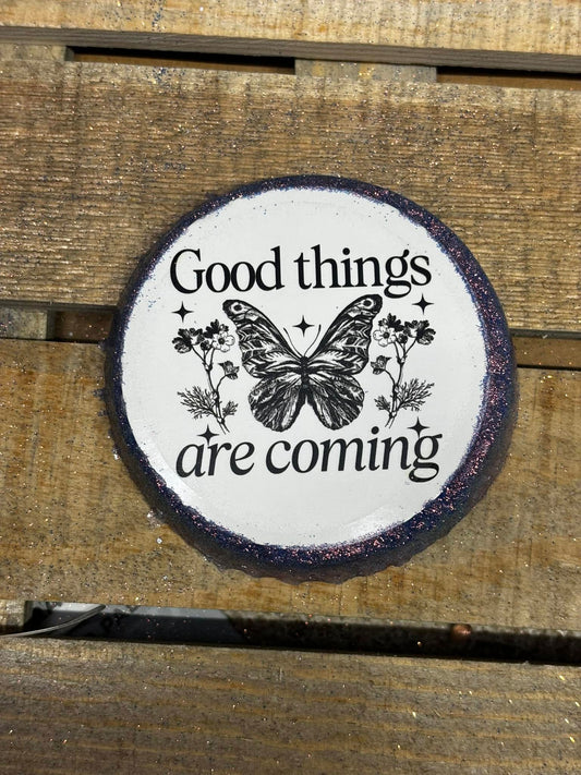 Round handmade freshie with black text that reads “Good things are coming,” centered around a butterfly and floral design on a wood background.