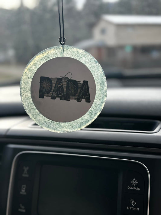 Handmade car freshie with cracked clear base and “PAPA” lettering filled with outdoor graphics, hanging in front of a car dashboard.