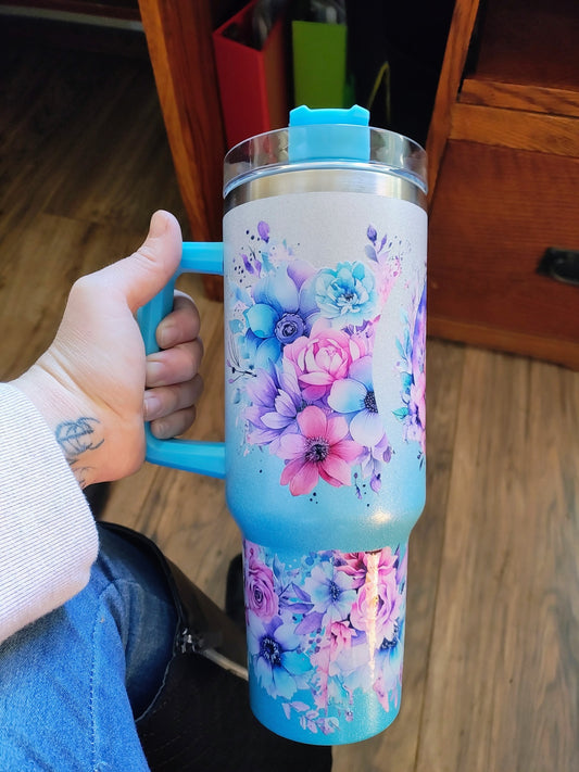 Blue 40oz handled tumbler with glitter finish and floral wrap design in pink, purple, and turquoise, held in hand over a wood floor.

