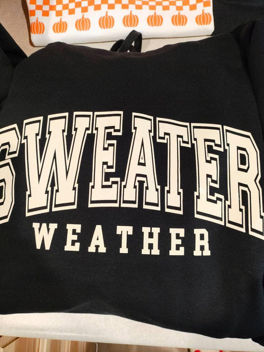 Black sweatshirt with large bold cream lettering that reads “Sweater Weather,” folded flat on a display table.

