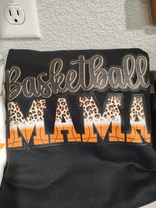 Black sweatshirt with “Basketball Mama” design in leopard and basketball print, folded on a shelf.

