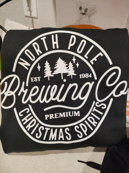 Black sweatshirt with white vintage-style “North Pole Brewing Co. – Christmas Spirits” graphic folded on a countertop.