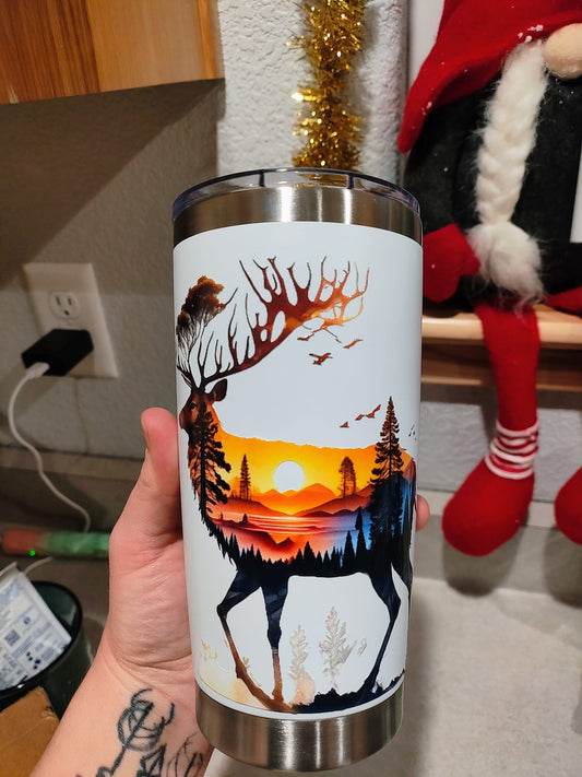 20oz stainless steel tumbler featuring an elk silhouette filled with a vibrant mountain sunset and pine tree landscape, held in a hand.

