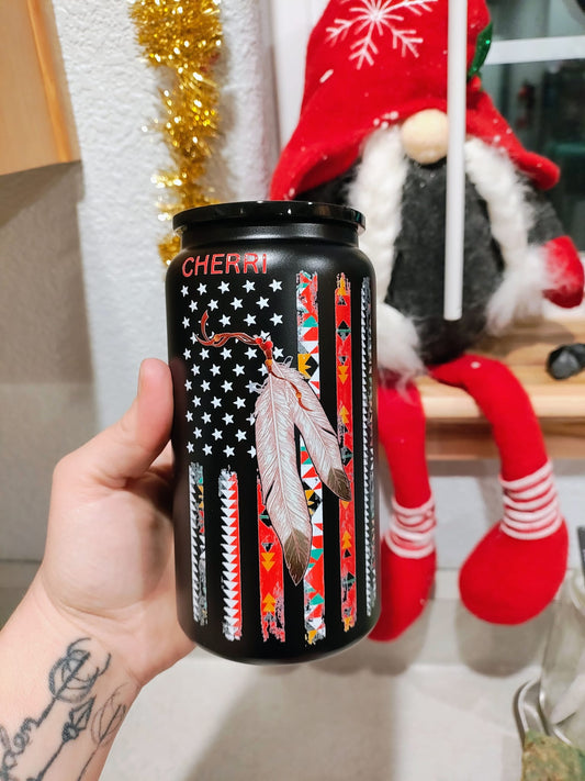Black can-shaped tumbler with tribal-patterned American flag, feathers, and the name “Cherri” on top, held in hand near a festive holiday background.