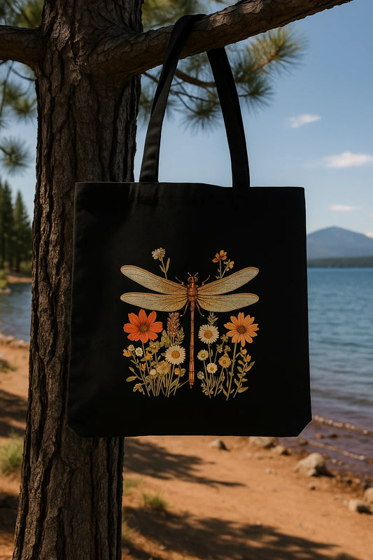 Black canvas tote bag with a printed gold dragonfly and wildflowers, hanging on a pine tree near a lake.