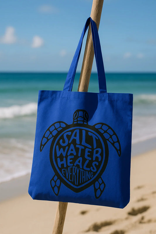 Blue canvas tote bag with a black sea turtle design and the words “Salt Water Heals Everything,” hanging on a stick at the beach.
