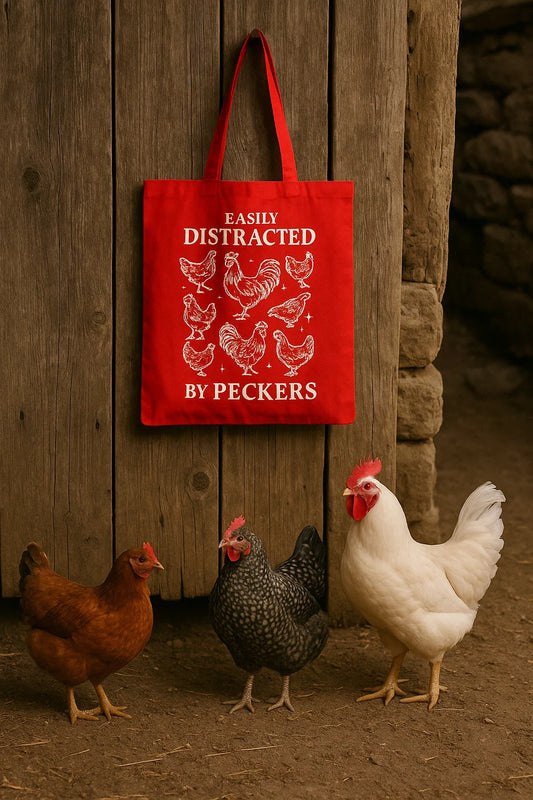 Red canvas tote with white chicken graphics and the text “Easily Distracted by Peckers,” hanging on a barn wall above real chickens.
