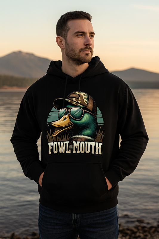 Man wearing a black hoodie with a duck graphic and 'Fowl Mouth' text, standing by a lake with mountains in the background.